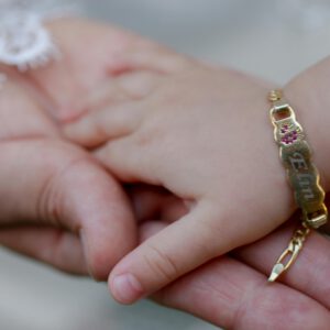 Close-up of a baby holding an adult's hand, featuring a gold bracelet with engraved name.